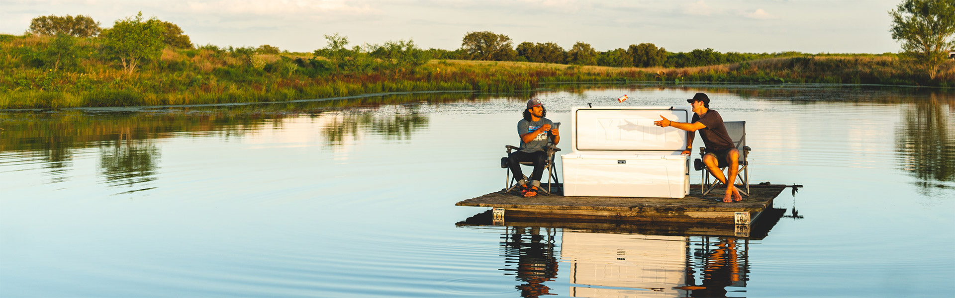 Two people on a small pontoon with a large YETI cooler,  on a calm lake with a scenic background