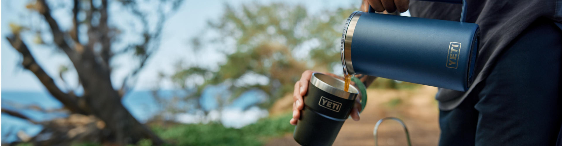 Person pouring coffee from a navy yeti french press  into a black tumbler outdoors with a scenic background.