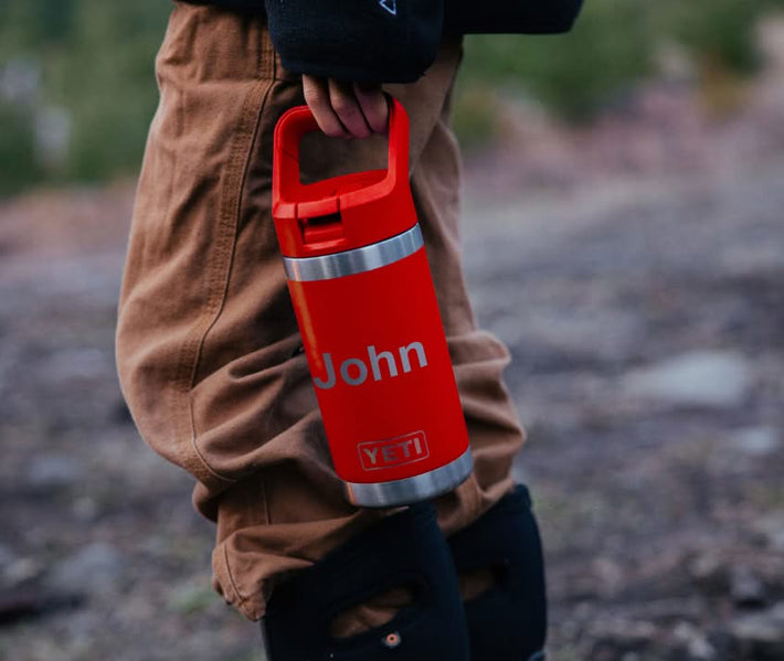 Person holding a red YETI bottle with 'John' printed on it, wearing brown pants and black boots in an outdoor setting.