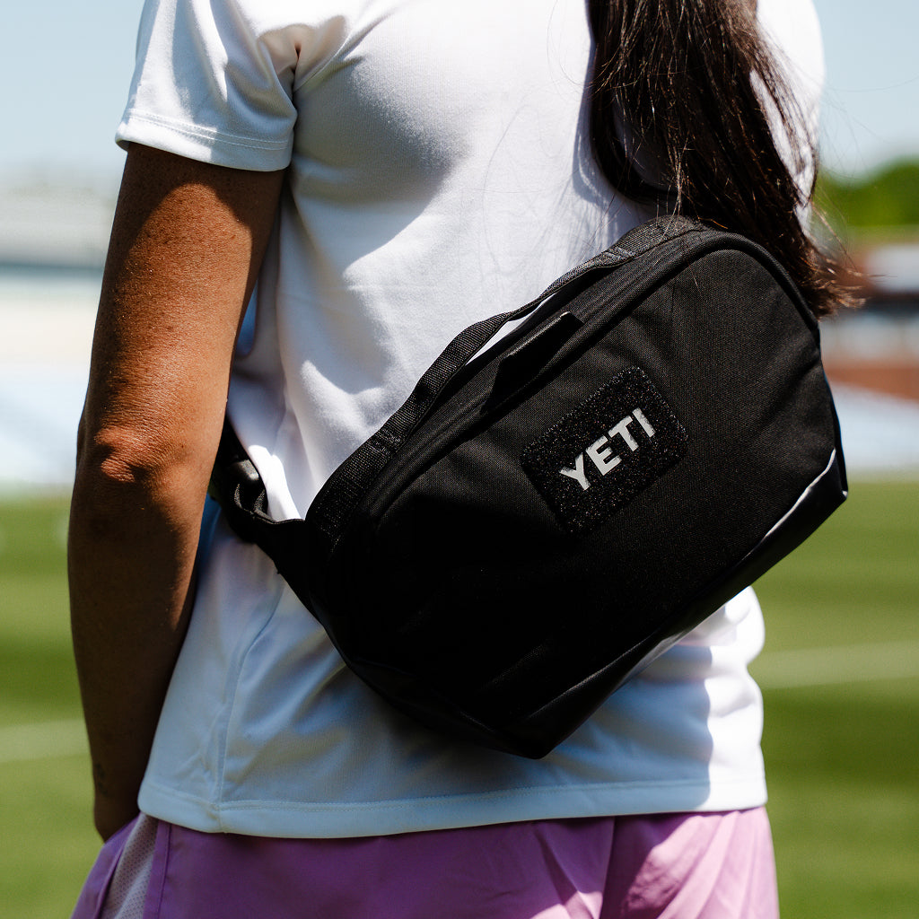 A person outdoors wearing a black YETI crossbody bag across their torso, with a white shirt and pink skirt visible against a blurred green lawn background.