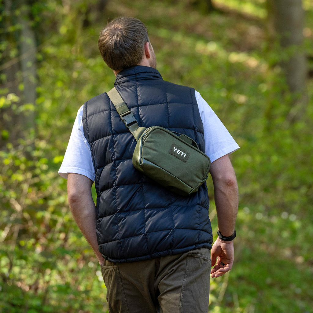 A man wearing a navy quilted puffer vest over a white t-shirt and olive pants, shown from behind in a forest while wearing an olive green YETI crossbody bag across his back.