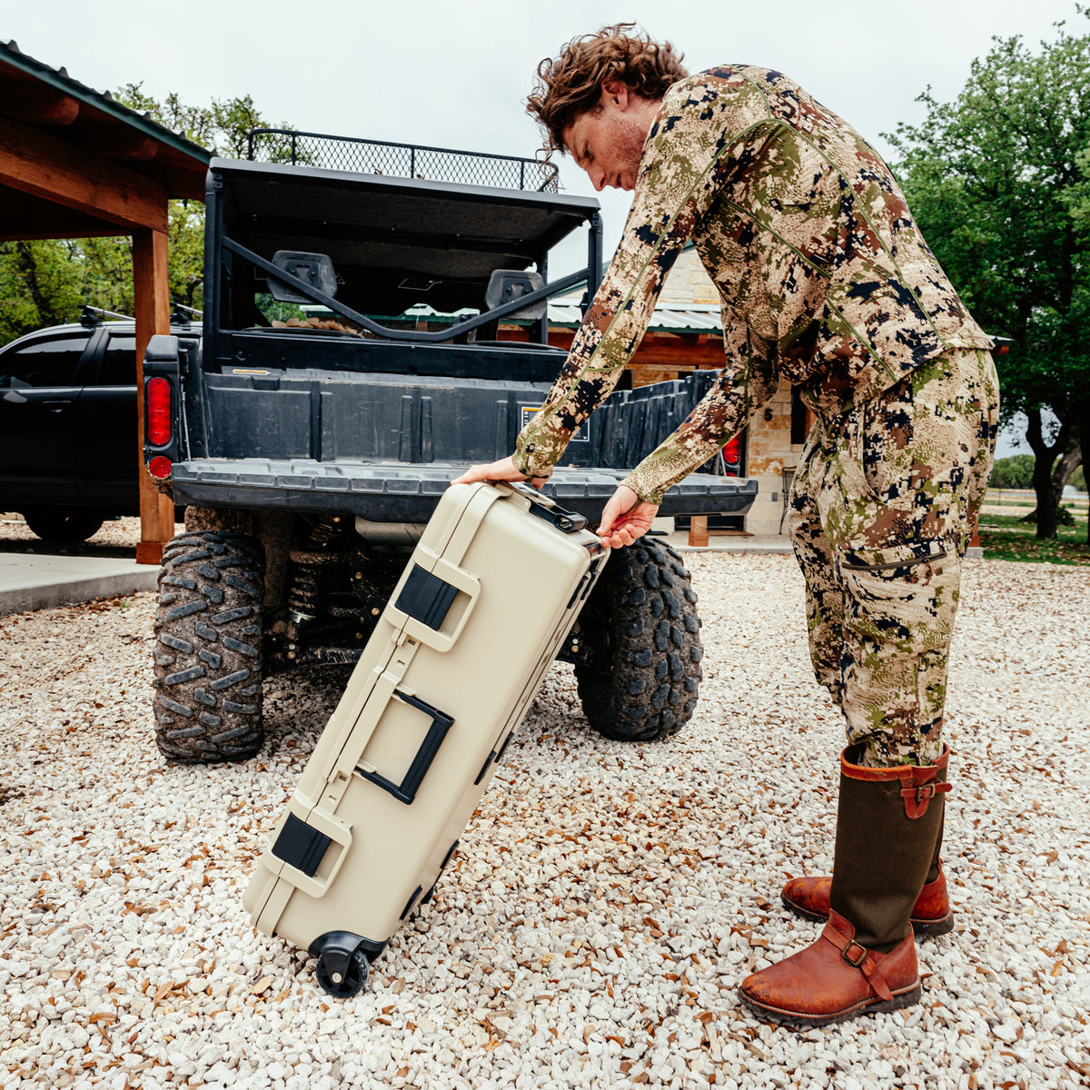 A man in a desert camouflage hunting suit and brown boots pushes a beige hard-shell rolling suitcase on a gravel driveway next to a black off-road truck.