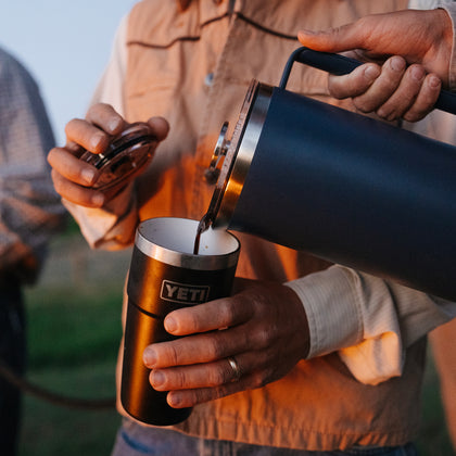 A person outdoors at sunset pours dark coffee from a matte black YETI insulated carafe into a matching black YETI tumbler. The warm golden hour light highlights the YETI branding on both vessels, with a blurred natural background.